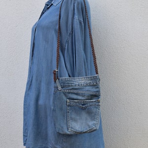 Puede incluir: Un bolso bandolera de mezclilla azul con una correa trenzada marr&oacute;n. El bolso tiene un cierre de solapa y un bolsillo frontal.