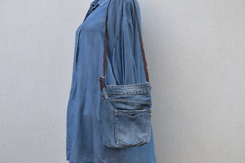 Puede incluir: Un bolso bandolera de mezclilla azul con una correa trenzada marr&oacute;n. El bolso tiene un cierre de solapa y un bolsillo frontal.