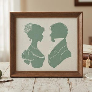 Op de afbeelding: Een ingelijst kruissteekkunstwerk met silhouetten van een man en een vrouw in profiel, in saliegroen. Het kunstwerk wordt weergegeven in een donkerhouten lijst, rustend op een lichtgekleurd houten oppervlak. Een kopje thee en andere decoratieve items staan op de achtergrond.