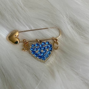 May include: Gold safety pin with a blue rhinestone heart charm and the letters 'S' and 'C' attached.