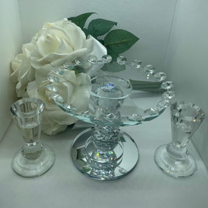 Puede incluir: Un soporte para tartas de cristal con una base de espejo y dos candelabros de cristal. El soporte está decorado con detalles de cristal y está rodeado de rosas blancas.