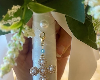May include: A close-up of a bridal bouquet handle wrapped in white satin ribbon. A gold charm with a cross design, adorned with pearls and a blue crystal, hangs from the handle. White flowers and greenery are visible in the background.