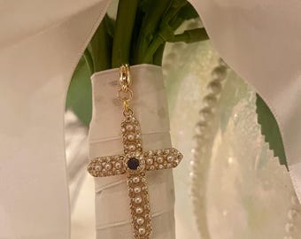 May include: A gold cross charm with a blue center stone and white pearl accents. The charm is attached to a white ribbon and is hanging from a bouquet of white flowers.