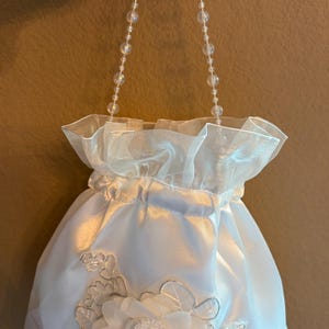 Puede incluir: Bolso blanco de satén con cordón y asa de cuentas. El bolso presenta una parte superior con volantes y una aplicación floral con detalles plateados. El asa está hecha de cuentas redondas y transparentes.