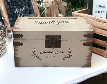 Peut inclure: Une boîte en bois rustique avec une finition blanche vieillie. La boîte est munie d'un loquet et de charnières en métal. La boîte est décorée des mots "Merci", "Laura & Luca" et "11 18 97".