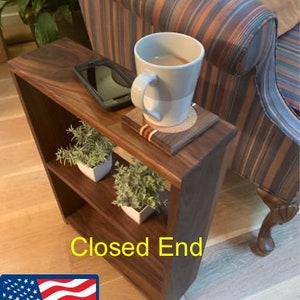 Puede incluir: Una mesa auxiliar de madera de nogal con un extremo cerrado, dos estantes y un portavasos. La mesa está junto a un sofá a rayas y tiene un teléfono y una taza encima. La mesa está etiquetada como "Closed End" y "Made in USA".