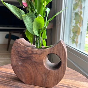 May include: A handcrafted wooden vase with a unique, curved design and a circular opening. The vase holds a small glass vial with pink and white flowers and green leaves. The vase is made of dark brown wood and sits on a wooden table.