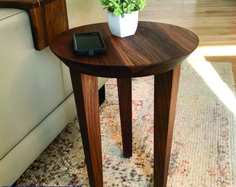 May include: A round dark brown wooden side table with three tapered legs. A small white square pot containing a green artificial plant and a black smartphone are placed on the tabletop. A matching wooden armrest tray is visible on the sofa nearby.