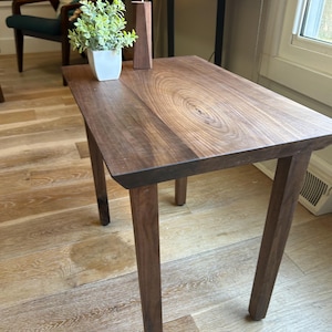 May include: A rectangular wooden side table with four legs. The tabletop features a natural wood grain pattern. A small white square pot with a green plant and a wooden candle holder are on the table.