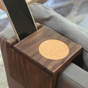 May include: A wooden phone and drink holder with a cork coaster. The holder is attached to the arm of a gray couch.