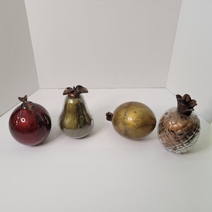Op de afbeelding: Vier decoratieve glazen fruitversieringen. Een rode granaatappel, een groene peer, een gouden ananas en een gouden en bruine eivormige versiering.