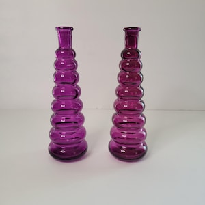 May include: Two purple glass vases with a bulbous, stacked design. The vases are tall and slender, with a rounded base.