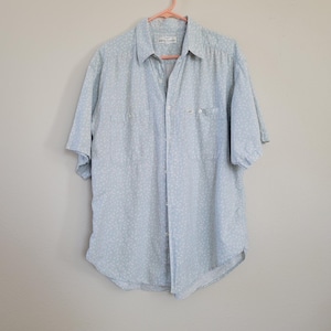May include: A light blue short-sleeved button-down shirt with a white floral pattern. The shirt has a button-up collar and two chest pockets.