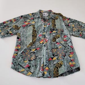 Puede incluir: Una camisa con botones y estampado. La camisa presenta un fondo verde azulado con formas geométricas rojas, amarillas y negras. Los diseños de remolinos negros y dorados contribuyen a su aspecto distintivo.