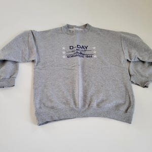 Könnte beinhalten: Graues Sweatshirt mit dem Text "D-DAY NORMANDIE 1944" in Marineblau. Das Design umfasst eine Flugzeuggrafik und Sterne. Das Sweatshirt hat einen Rundhalsausschnitt und lange Ärmel.