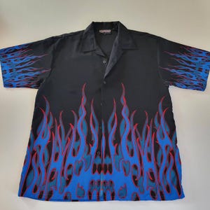 Puede incluir: Camisa negra con un diseño de llamas azules y rojas. Las llamas están impresas en la parte delantera, trasera y mangas. La camisa tiene cuello y mangas cortas. El diseño evoca la cultura de los hot rods.