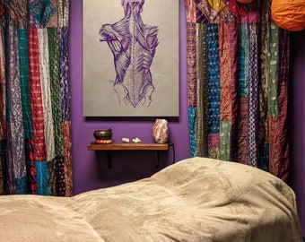 May include: A massage table with a beige sheet, a purple wall, and a vintage anatomical illustration of the back muscles. The room is decorated with colorful curtains and a small wooden shelf with a bowl and a salt lamp.