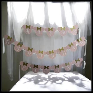 Puede incluir: Bandera rosa con la frase "HAPPY BIRTHDAY" escrita con letras y lazos dorados con purpurina. La bandera tiene forma de cabezas de Mickey Mouse. Se adjuntan cintas blancas para colgarla. Se muestra frente a una cortina blanca.