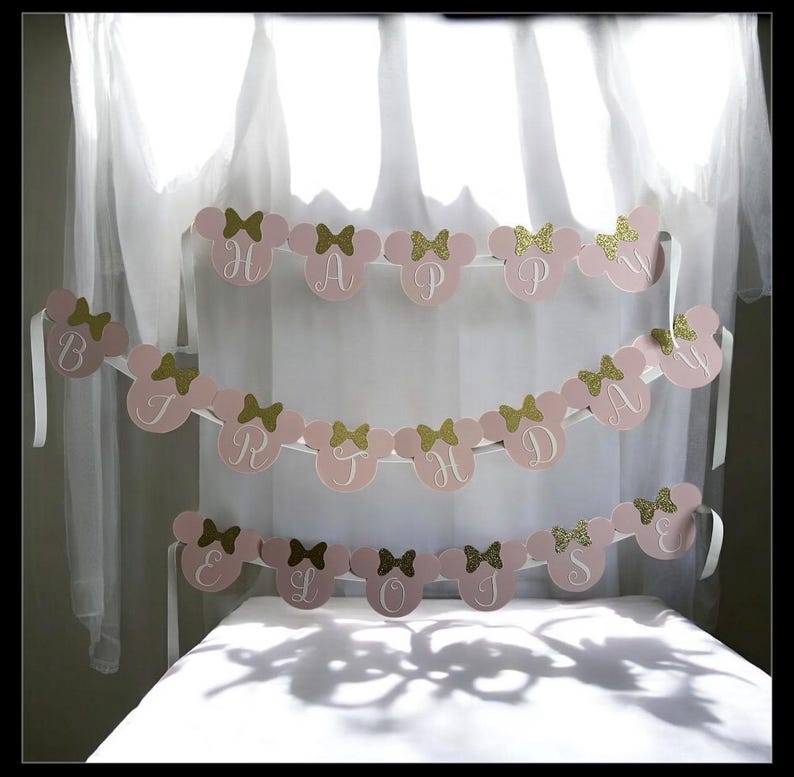 Puede incluir: Bandera rosa con la frase "HAPPY BIRTHDAY" escrita con letras y lazos dorados con purpurina. La bandera tiene forma de cabezas de Mickey Mouse. Se adjuntan cintas blancas para colgarla. Se muestra frente a una cortina blanca.