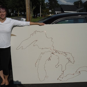 May include: A white canvas with a copper wire outline of the Great Lakes. A woman in a white shirt and black trousers is holding the canvas.