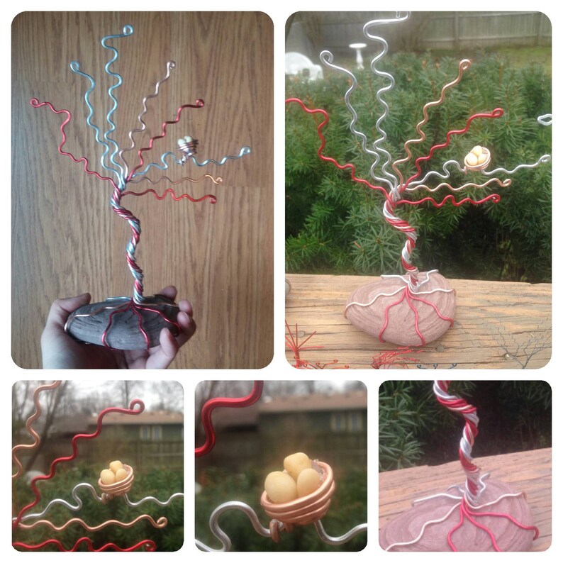 May include: A wire sculpture of a tree with red, silver, and copper wire branches. The tree is standing on a rock base and has a small nest with three white eggs in one of the branches.