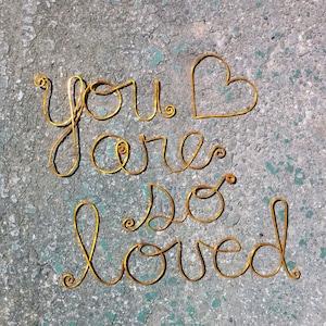 Puede incluir: Escultura de alambre dorado sobre un fondo gris que dice "You are so loved" con una forma de corazón en la palabra "you".