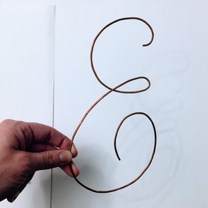 May include: A copper wire shaped into the letter "E" with a decorative, cursive style.