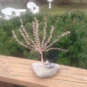 May include: A wire sculpture of a tree with a copper-colored trunk and branches. The tree is sitting on a gray rock with a smaller blue wire sculpture of a tree on top of it.