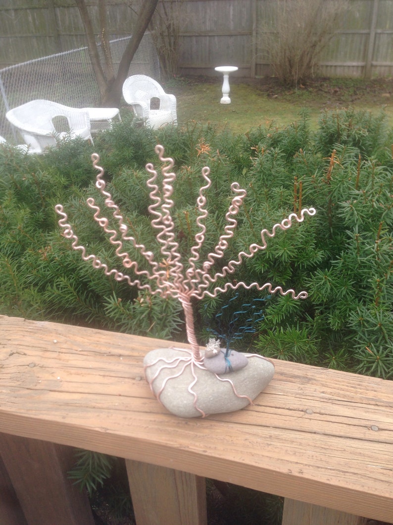 May include: A wire sculpture of a tree with a copper-colored trunk and branches. The tree is sitting on a gray rock with a smaller blue wire sculpture of a tree on top of it.