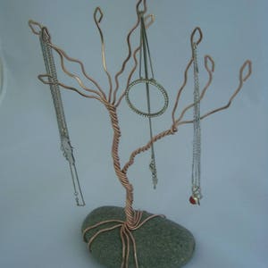 May include: A copper wire jewelry tree with a gray stone base. The tree has several branches that are bent into hooks to hold necklaces and earrings. There are several necklaces and earrings hanging from the tree.