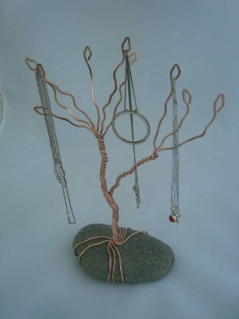 May include: A copper wire jewelry tree with a gray stone base. The tree has several branches that are bent into hooks to hold necklaces and earrings. There are several necklaces and earrings hanging from the tree.