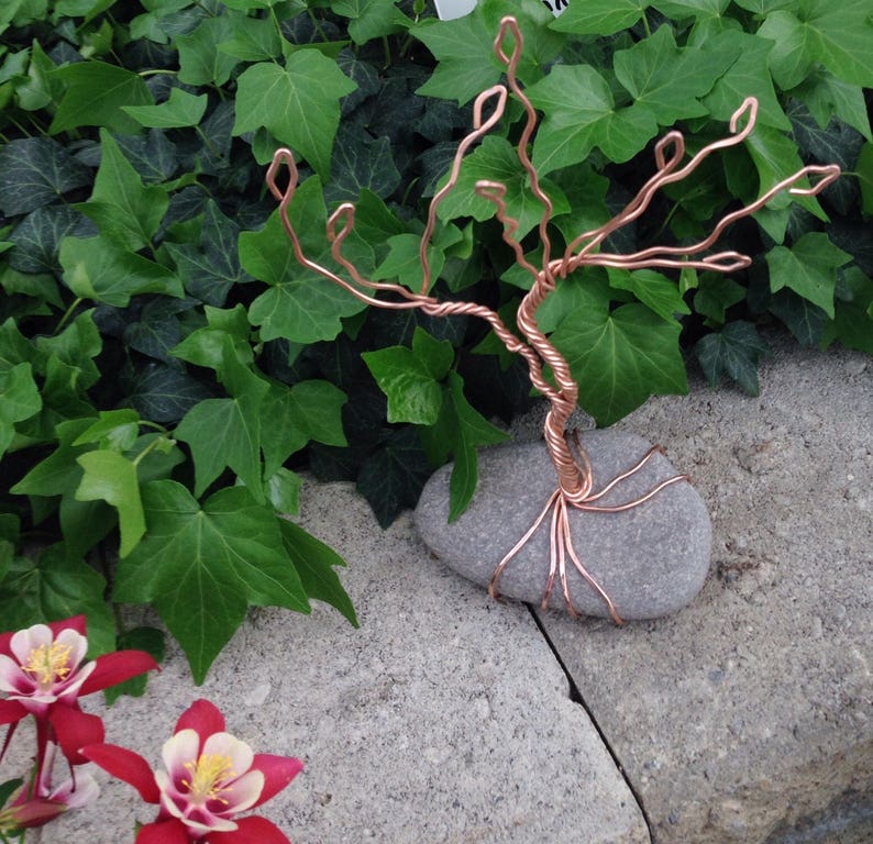 May include: A copper wire tree sculpture with a twisted trunk and branches, set on a gray stone base. The sculpture is placed on a bed of green ivy.