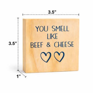 May include: A wooden block, 3.5 inches square and 1 inch thick, with the text "YOU SMELL LIKE BEEF & CHEESE" in dark blue. Two blue heart shapes are below the text. The block is made of light-colored wood.