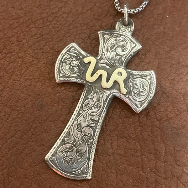 May include: A silver cross pendant with intricate floral engraving and a gold 'WR' monogram. The pendant is on a silver chain.
