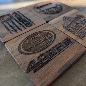 Puede incluir: Cuatro posavasos cuadrados de madera con diseños grabados. Un posavasos presenta el logotipo y el texto de los 49ers, otro tiene un diseño de puente con texto y los otros tienen otros diseños. Los posavasos son de madera oscura.