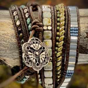 May include: A multi-layered leather wrap bracelet with a silver charm. The bracelet features various beads in shades of green, brown, and white. The charm is a silver flower with a triangular design in the center. The bracelet is wrapped around a piece of wood. The text "Ginger Blossom Studios" is visible at the bottom of the image.