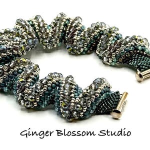May include: A beaded bracelet with a spiral design. The bracelet features a mix of teal, silver, and clear beads. The clasp is silver. The text "Ginger Blossom Studio" is at the bottom.