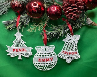 May include: Three white lace Christmas ornaments with red embroidered names: Pearl, Emmy, and Tripod. The ornaments are hanging from a green background with red berries and a pine cone.