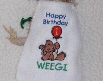 May include: A bearded dragon wearing a white bib and party hat. The bib has the text "Happy Birthday 1" with a cartoon bear holding a balloon and the name "WEEGI". The hat is white with green polka dots.