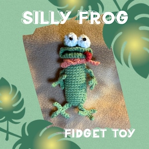 Pode incluir: Um brinquedo fidget de crochê de sapo verde com olhos brancos, um cachecol rosa e uma língua vermelha. O texto "SILLY FROG" e "FIDGET TOY" são exibidos em branco. O fundo tem um esquema de cores verde e marrom.