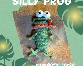 May include: A green crocheted frog fidget toy with white eyes, a pink scarf, and a red tongue. The text "SILLY FROG" and "FIDGET TOY" are displayed in white. The background has a green and brown color scheme.