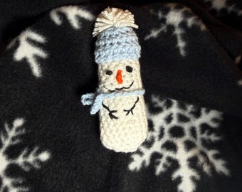 May include: A handmade crochet snowman with a light blue hat and scarf. The snowman has a carrot nose, black eyes, and a smiling mouth. The snowman is on a black background with white snowflake patterns.