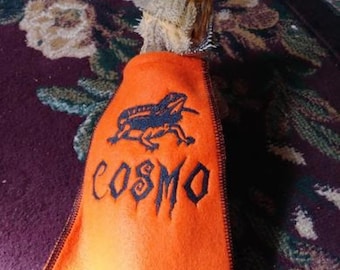 May include: An orange cape with black trim and the name "COSMO" embroidered in black, along with a black lizard graphic. The cape is worn by a bearded dragon, with the background showing a patterned rug.