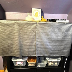 Peut inclure: Une housse en tissu gris foncé rectangulaire avec un motif géométrique répétitif drapée sur une étagère noire. La surface supérieure contient une figurine blanche, une carte rectangulaire avec une grenouille jaune et le texte PERTY.