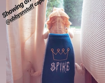 May include: A bearded dragon wearing a blue cape with a gold crown and the name "SPIKE" in white script. The image includes the text "SUPER SPIKE" and "#BEARDEDDRAGONS". The reptile is looking out a window.