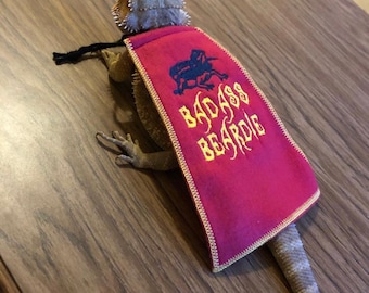 May include: A bearded dragon wearing a red cape with yellow embroidered text that reads "BADASS BEARDIE". The cape also features a black and blue embroidered emblem. The cape is trimmed with a white stitched border.