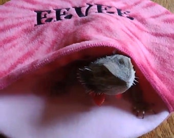 Pode incluir: Uma cama de animal de estimação de tecido listrado rosa e branco com a palavra "EVER" bordada. Um dragão barbudo está parcialmente escondido dentro da cama.