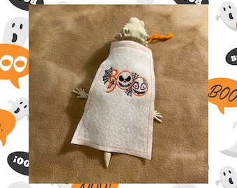 May include: A white felt ghost decoration with an embroidered "Boo" design featuring a skull and two cartoon faces. The ghost has small arms and a wooden stake at the bottom. An orange string is attached to the top.