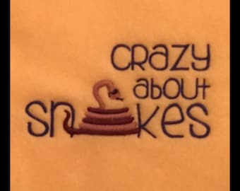 May include: Embroidered design on an orange background. The text reads "Crazy about Snakes" in dark blue script. A brown and tan snake is coiled below the text, adding a playful touch to the design.