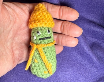 May include: A handmade, green crocheted figure with a yellow hat and scarf. The figure has a simple face with black eyes and a mouth, and white tear drops. The figure is held in a hand against a purple background.
